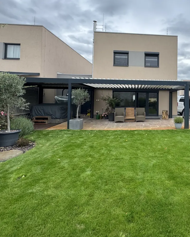Stejný dům dnes: dvě tmavé pergoly s lamelami, carport a zahrada s trávníkem.