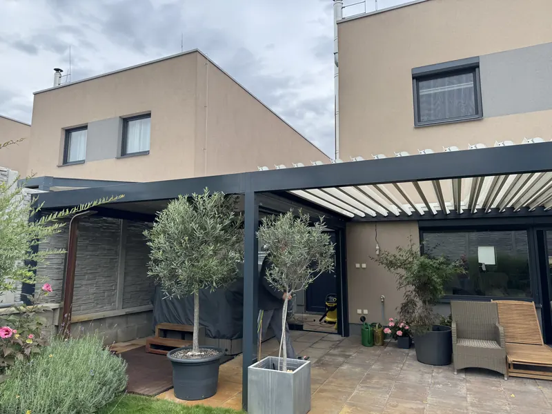 Pergola u domu s olivovníky v květináčích a posezením.